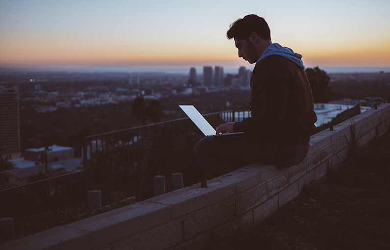 man sitting on a rooftop with a laptop, with a stunning city view in the sundown