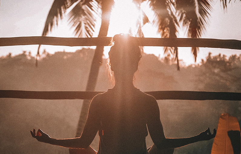 woman seen from behing sitting in lotus position with a view of a palm tree and the sun shining